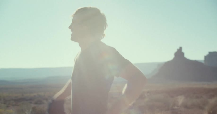 A man with long curly brown hair takes off his black jacket and throws it over his shoulder while standing in a wide desert landscape he is silhouetted against the bright sunlight as he turns and begins to jog away