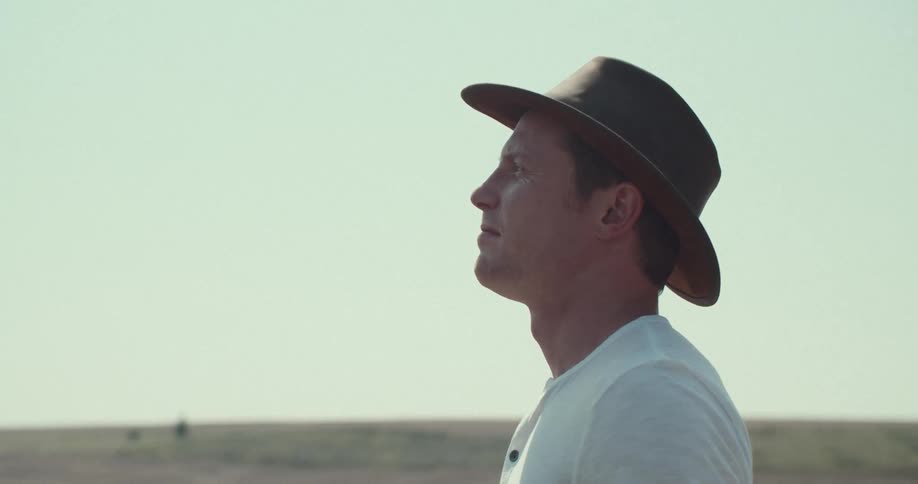 A lowangle shot shows a man in a white tshirt jeans and a cowboy hat standing on a dirt mound hes facing away from the camera gazing out at the vast desolate landscape