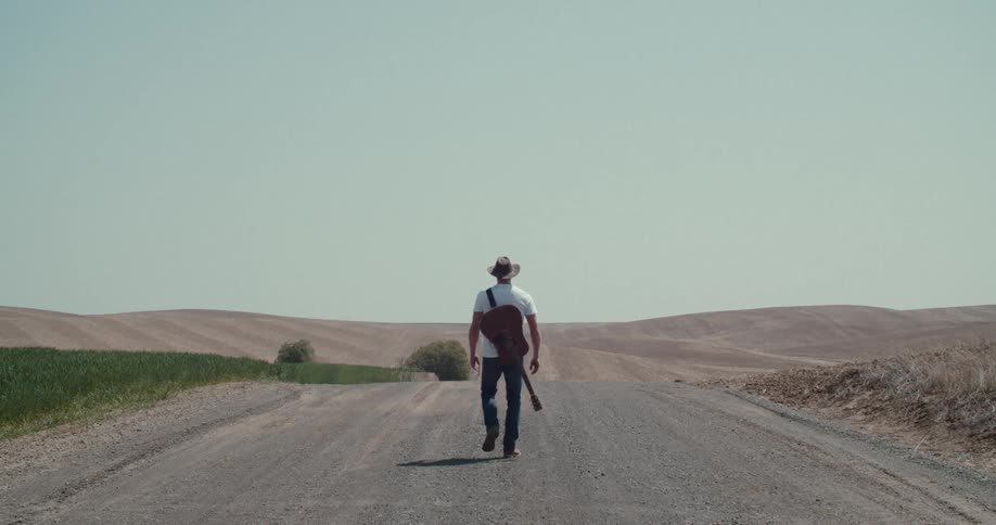A man with a brown hat walks down the middle of a dirt road with a guitar strapped to his back while the road passes through a valley of low hills and fields under a wide pale sky