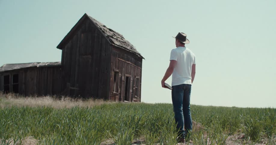 A male wearing a hat stands in tall grass looking toward an old weathered wooden house
