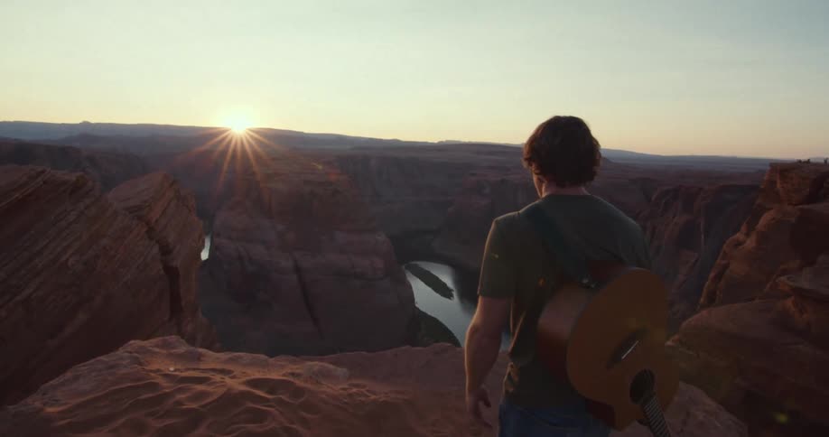 A young man walks along a canyon cliff at sunset carrying a guitar on his back