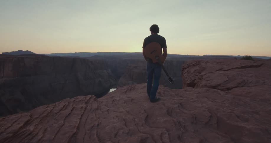 A solitary figure with a guitar stands atop a vast cliff overlooking an expansive canyon at sunset