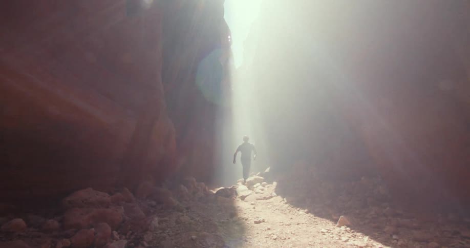 A wide view shows a person wearing a dark shirt and jeans walking through a rocky slot canyon as a bright beam of light shines down through the narrow opening above