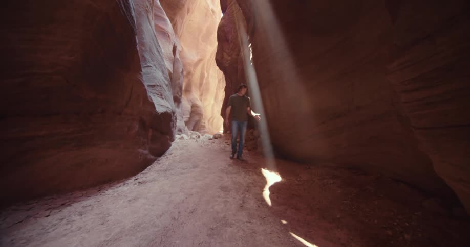 A man walks along a narrow dirt path inside a tall slot canyon with a bright beam of sunlight shining down onto the path in front of him