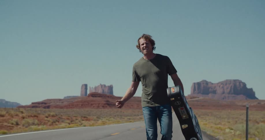 A man with a guitar case walks down the middle of a long paved road in a vast desert landscape with a red rock mountain range in the distance