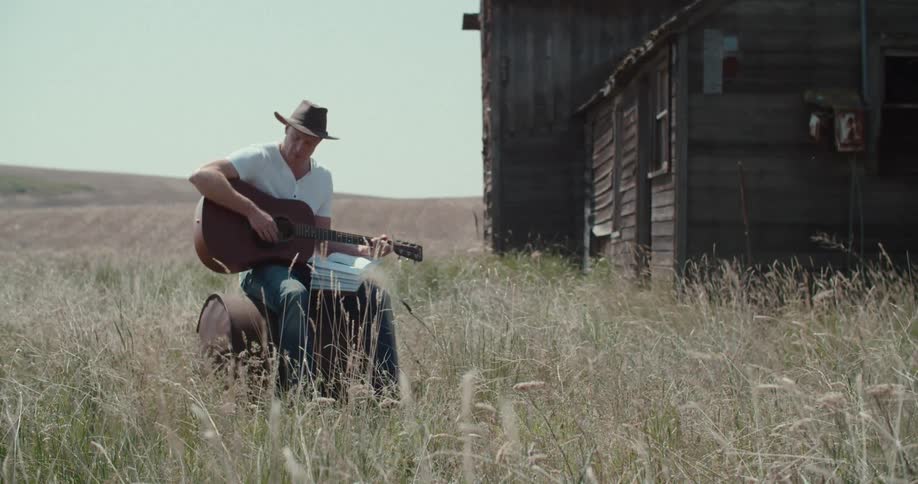 A man wearing a cowboy hat jeans and a white tshirt sits on a rusted barrel in a field of tall dry grass hes playing an acoustic guitar while looking at a music book on his lap with a weathered wooden cabin visible behind him