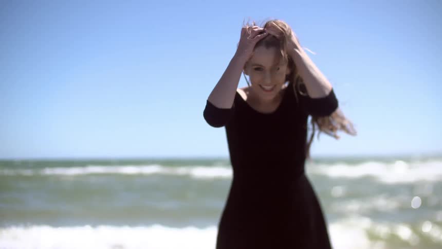 A young, blonde woman in a black dress smiling and brushing her wind blown hair back from her face as waves roll on shore behind her