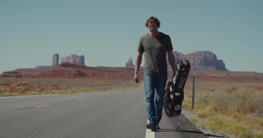 A man with a guitar case walks along the shoulder of a desert highway he is looking for a ride and raises his thumb as a car drives by in the background