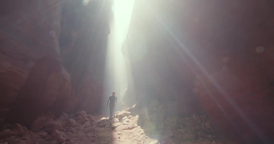The video shows a person standing in a narrow slot canyon with a beam of sunlight shining down the two lines are the parallel walls of the canyon which create two distinct almost vertical lines framing the person and the bright light