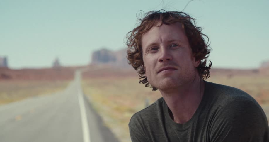 A man with curly brown hair sits on the side of a lonely desert road he looks back over his shoulder at the camera with a serious and contemplative expression