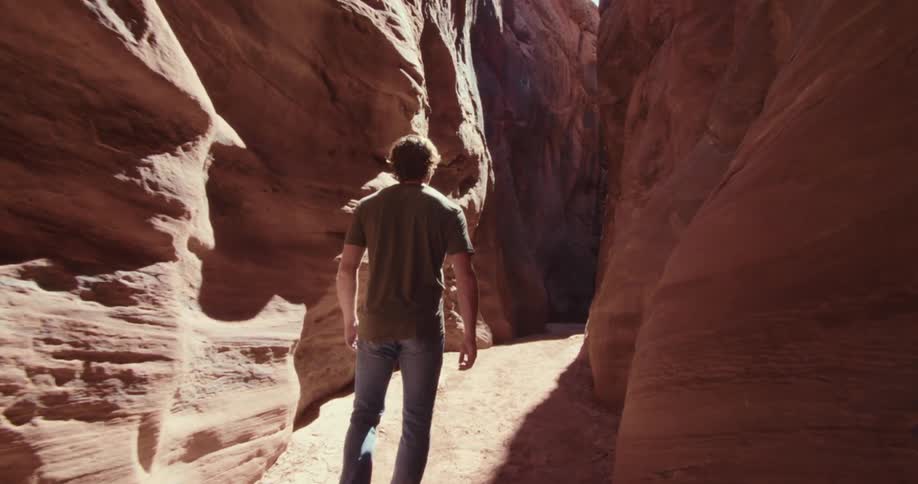 A man wearing a gray shirt and jeans walks away from the camera and stops to look up and reach out with one hand to touch the side of a tall light brown slot canyon