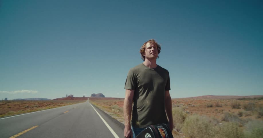 A male walks down a desert road carrying a musical instrument case under a clear sky