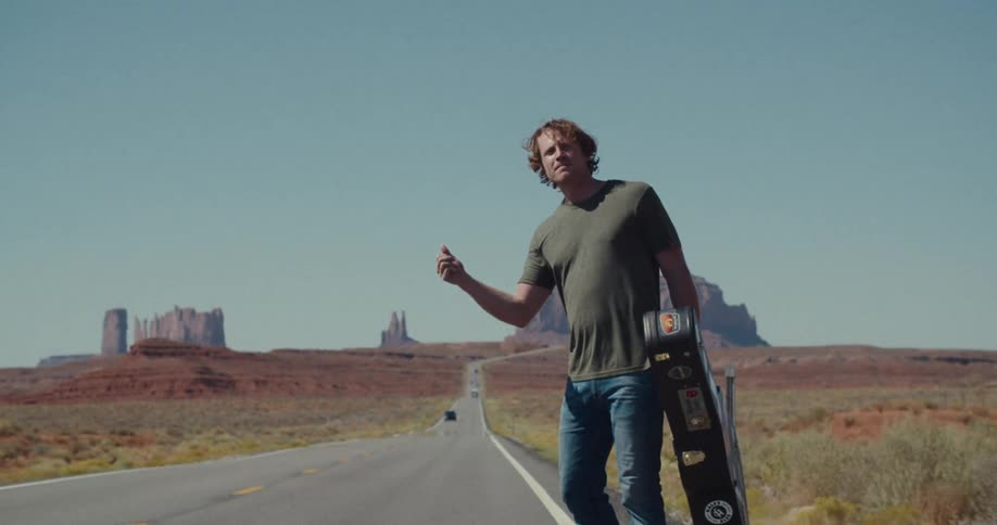 A male stands on a desert highway holding a guitar case and gesturing with his thumb as if hitchhiking