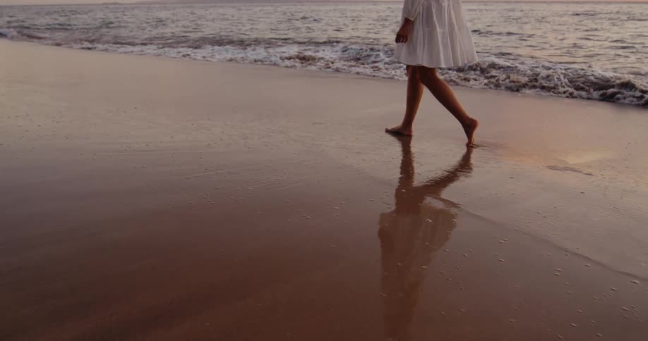A female in a white dress walks barefoot along the shoreline with her reflection visible on the wet sand