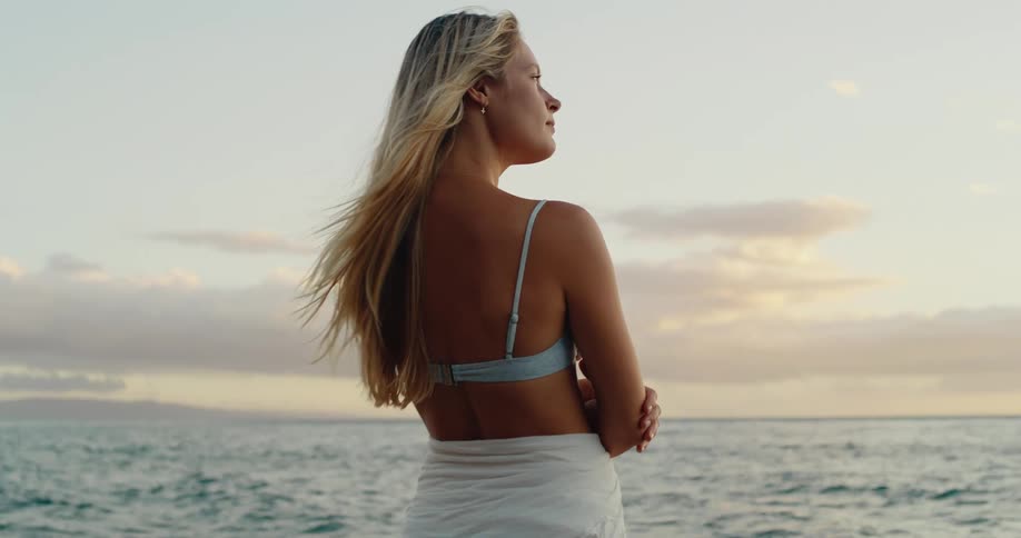A female with long blonde hair stands near the ocean looking toward the horizon during sunset