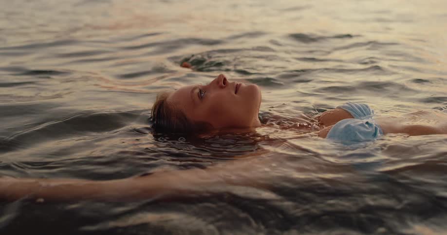 A young woman floats on her back in the ocean as the sun sets she wears a light blue bikini top with her arms stretched out to the sides and her eyes closed in peaceful tranquility
