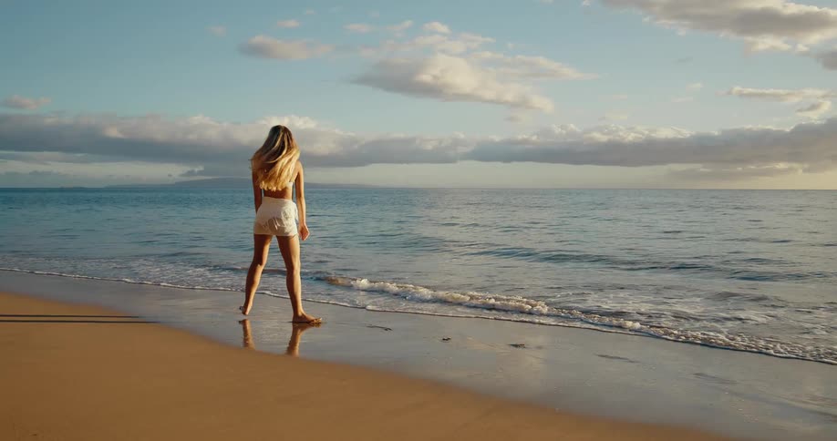 A person walks on a beach at sunset with waves on one side and a reflection of the sky on the other