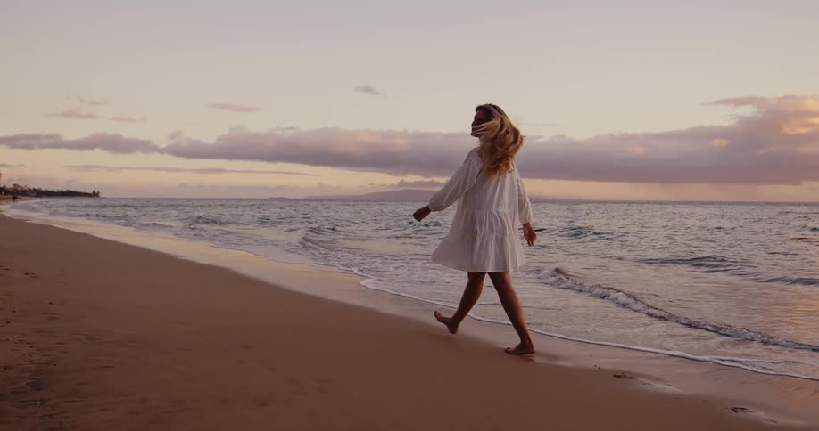 A woman walks on the sand at the edge of the water and turns her body toward the water