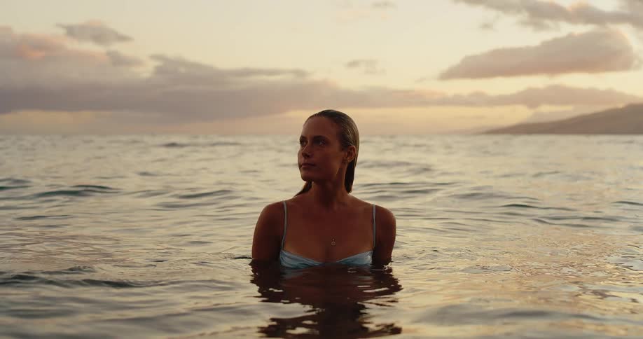 A young woman wearing a swimsuit stands in the ocean with her waist submerged at sunset