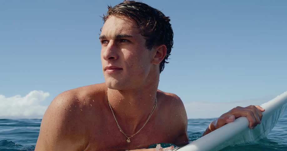 A male surfer paddles on his surfboard in the ocean looking around and then directly at the camera
