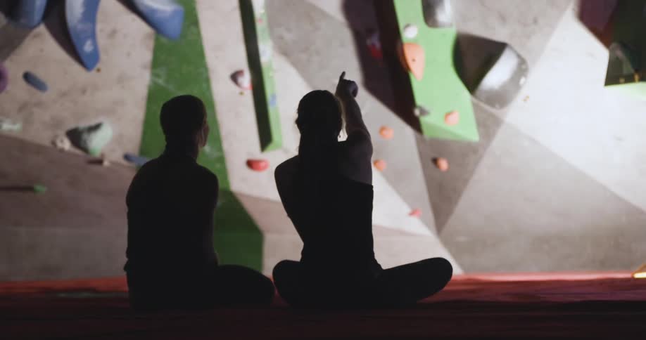 The video shows the silhouettes of two people sitting on the floor in a rock climbing gym one person points up toward a climbing route on the wall as they talk to the other person who is seated beside them both are discussing the route before they get