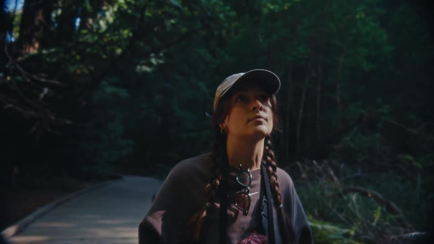 A video shows a young woman with brown hair in two braids wearing a baseball cap and sunglasses around her neck she is in a forest and looks up seemingly captivated by something she sees