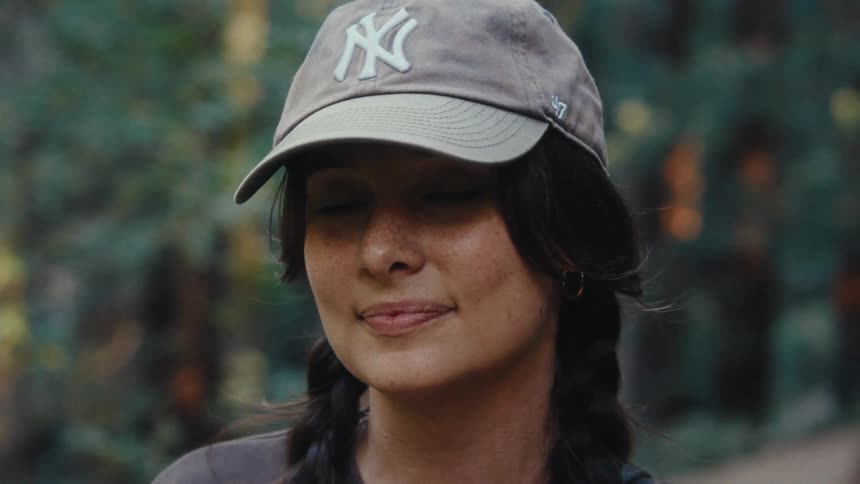 The first line shows a young woman with a baseball cap on and braided hair smiling in a forested area the second line is a slow soft zoom into a tight shot on her face showing her freckles and a gentle smile