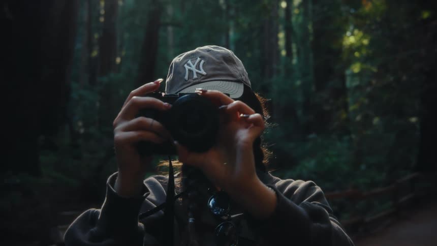 The video shows a person wearing a grey new york yankees baseball cap and a grey hoodie the person is holding a camera up to their face obscured by the camera and seems to be taking a photo in a forest after a few seconds they slowly lower the camera