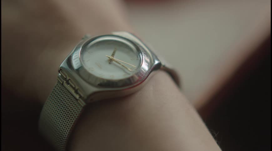 A video shows a closeup shot of a persons wrist wearing a modern silvertoned watch with a simple white face and a mesh band the camera focuses on the watch as the light catches the metallic surface