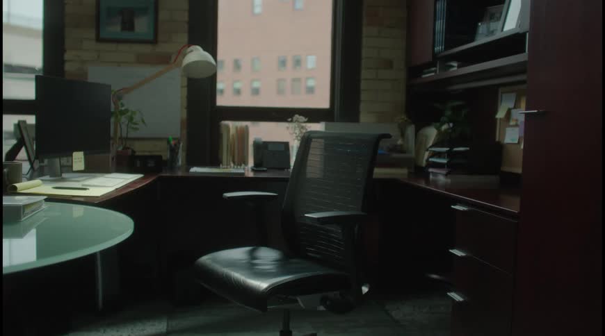 A well organized but empty office features a dark wood desk with a computer and a lamp an ergonomic black chair and a large window looking out onto a brick building