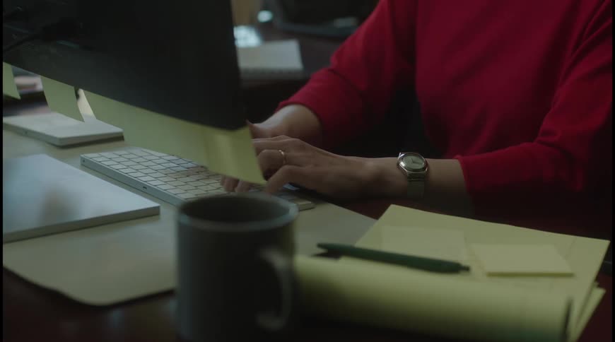 Person in red types on the keyboard at a desk with a mug notebook and notes
