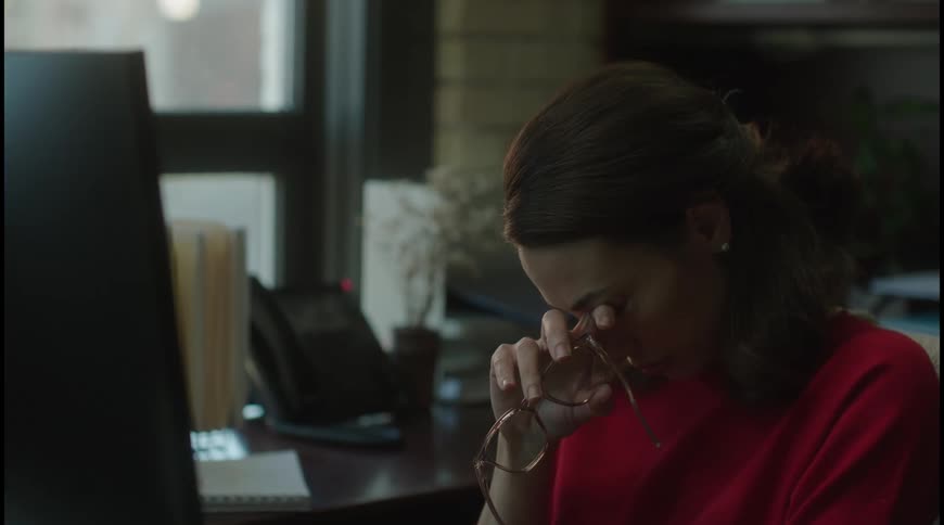 Woman in red holds her glasses and rests her face in a gesture of fatigue at the office