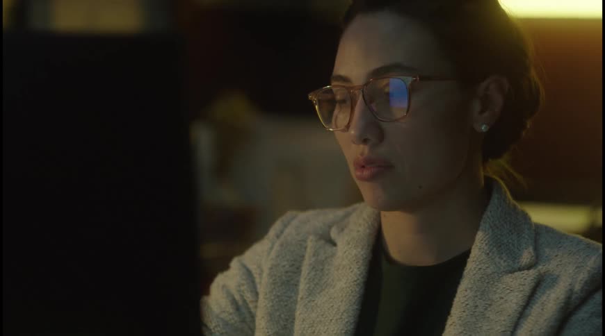 A woman wearing glasses leans forward while looking at a computer screen