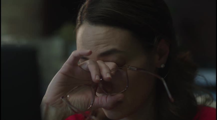 A young woman wearing a red dress typing on a computer takes off her glasses to wipe tears from her eyes