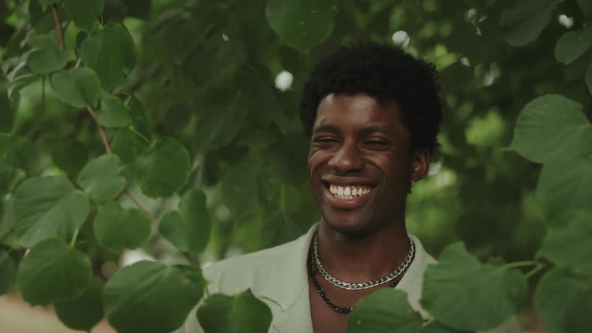 Portrait of a smiling young man among green branches wears a light shirt and necklaces