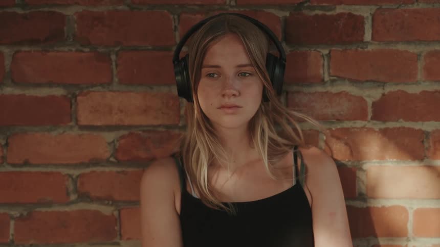 Young blonde woman wears black headphones leaning against a brick wall