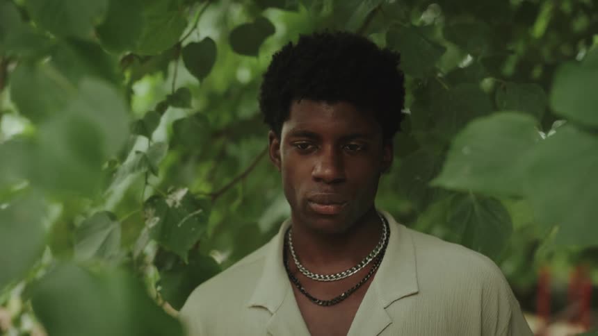 A man with a serious expression and a short curly afro looks down as he stands among green leaves wearing a white collared shirt and two necklaces