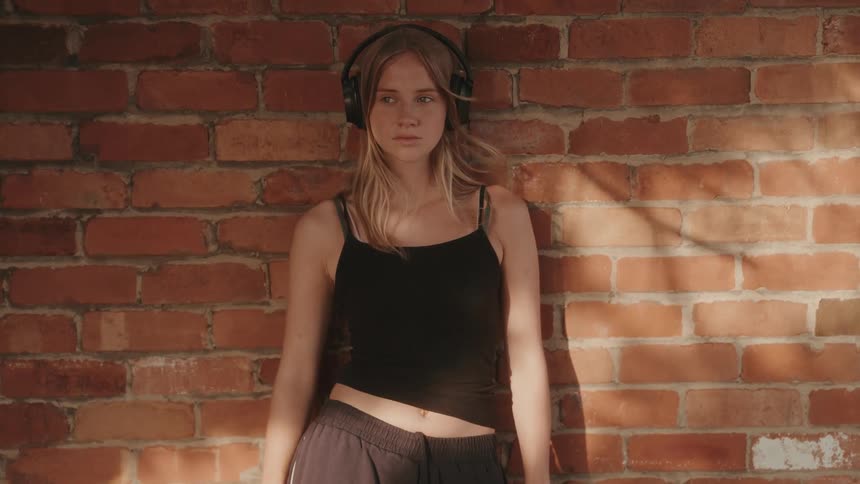 Young woman in black with headphones crosses her arms leaning against a brick wall