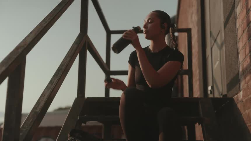 A young woman with a ponytail wearing a black t-shirt sits on an outdoor staircase drinking from a water can at sunset