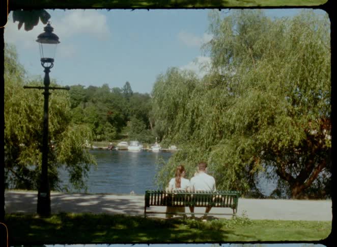 A couple sits on a bench overlooking a riverside with boats gliding on the water