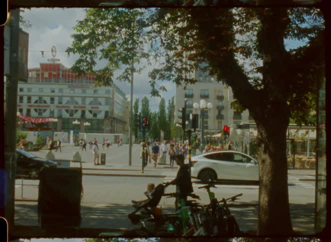 The video captures a lively city intersection with people walking and cars driving past the footage has a vintage film look framed by trees and showing film sprocket holes on the side