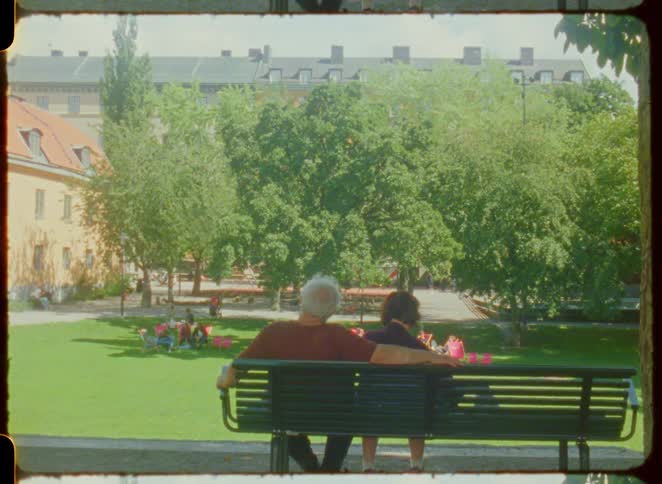 A couple sits closely on a park bench in a lush green park