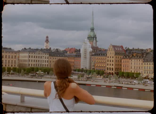A person stands on a bridge with their back to the camera and looks at a body of water and a city in the background