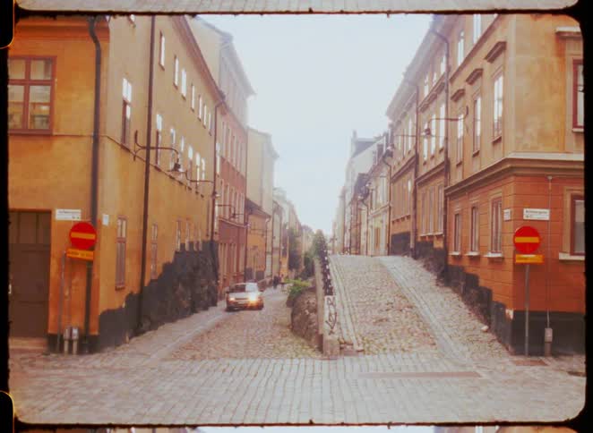 Car drives along a cobblestone street between old buildings in orange tones