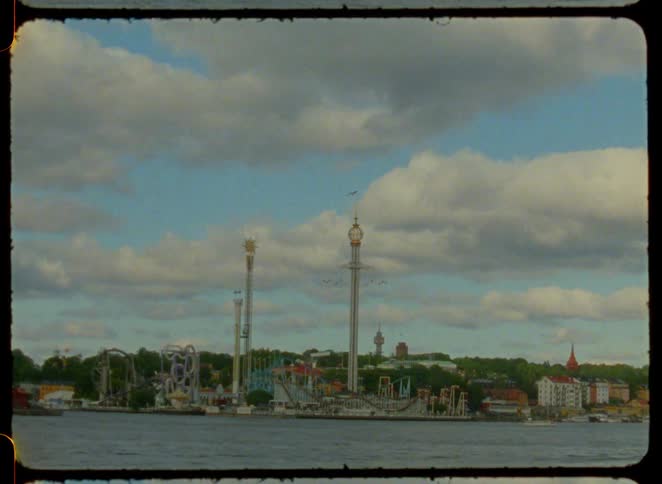 A full view from the water shows a city amusement park on the shore with roller coasters and tall drop rides and a small boat passes a flying seagull