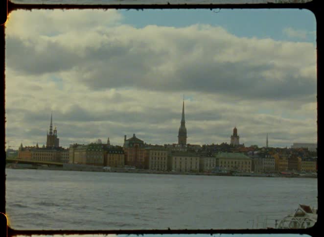 A tour boat gliding across a body of water in front of a historic european cityscape with prominent steeples under a cloudy sky