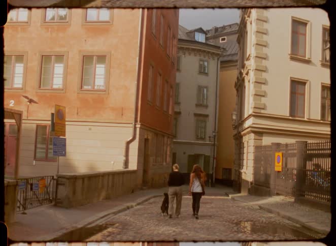 Two people and a black dog on a leash walk down a cobblestone street between buildings and a stone staircase a scene viewed through the tan ragged frame of a film strip