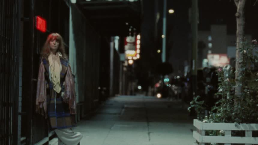 A woman stands on a dim sidewalk at night with city lights behind
