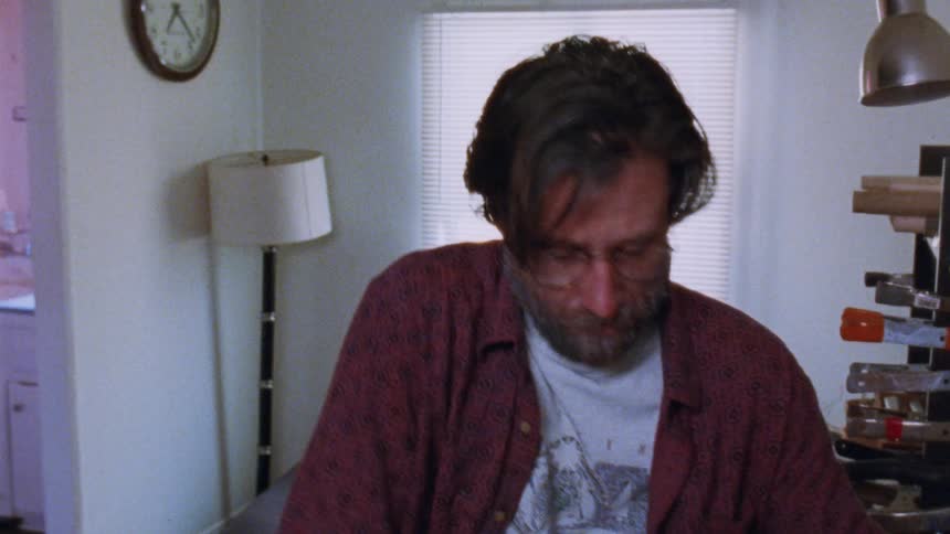 A man with a beard and glasses places his hand on the shoulder of a younger man with curly hair the younger man looks down seemingly in distress while the older man says something to him then turns and walks away with a duffel bag