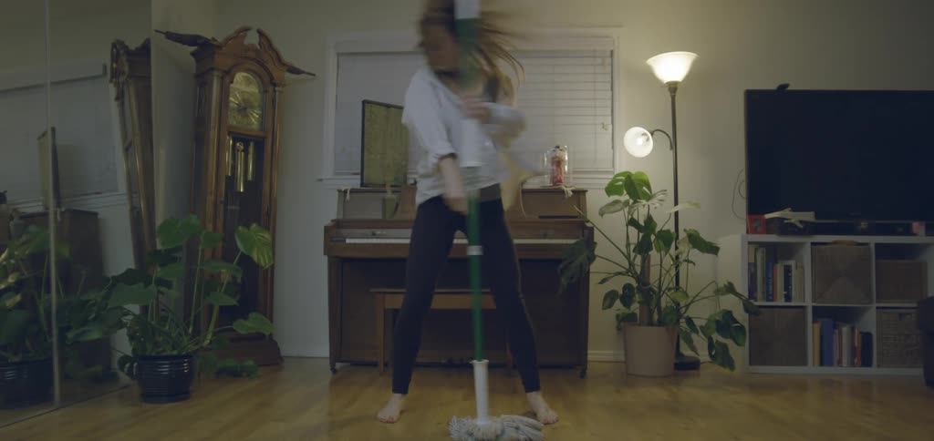 A woman smiles and dances joyfully while mopping the wooden floor of her home she moves with carefree energy using the mop as a prop in her lively solo dance routine